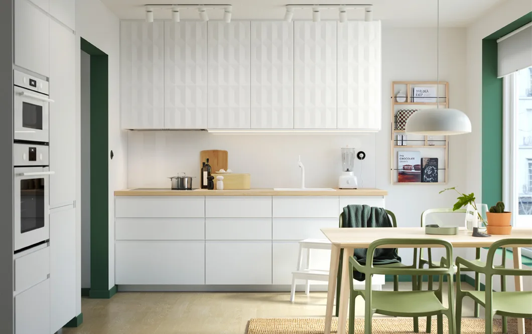 A white kitchen with green accents. Sleek white VOXTORP cabinet doors can be seen on the lower section and 3-dimensional HERRESTAD cabinet doors on the upper section. An LISABO table in ash veneer and YPPERLIG chairs in green can be seen in the foreground.